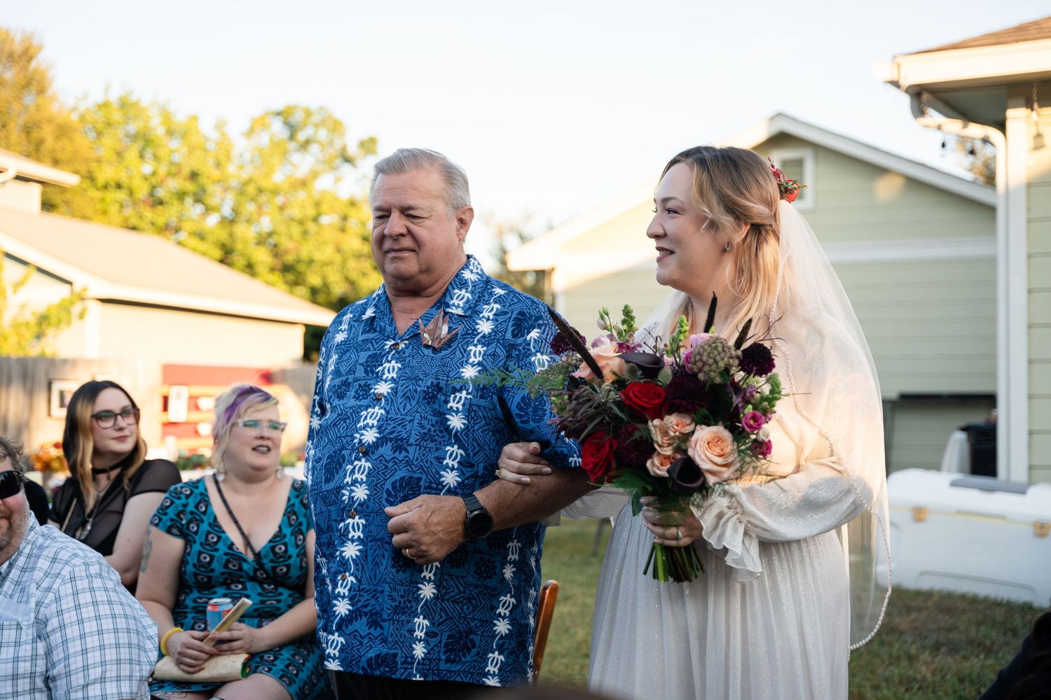 Wedding attendees gather outdoors as a bride holds a vibrant red and pink bouquet.