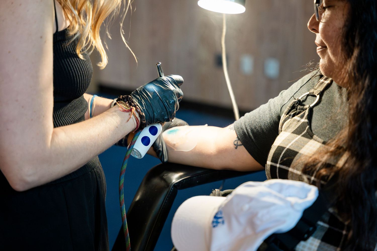 Tattoo artist wearing blue gloves works on a client's arm in a dimly lit studio under a lamp.