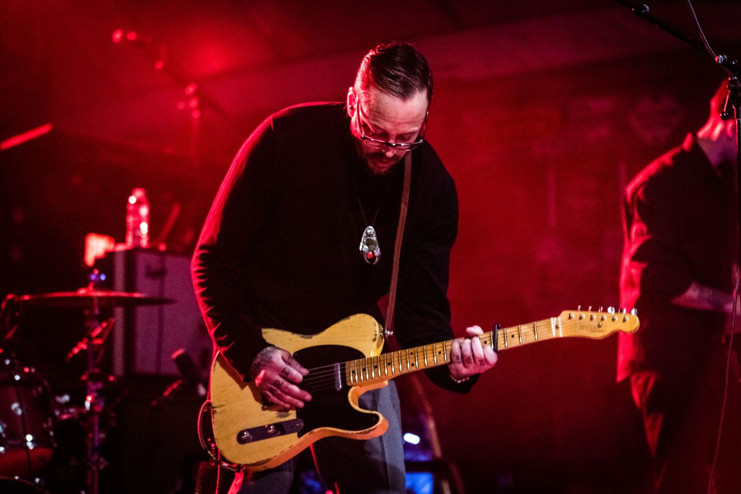 Guitarist performing with yellow Telecaster under dramatic red concert lighting.