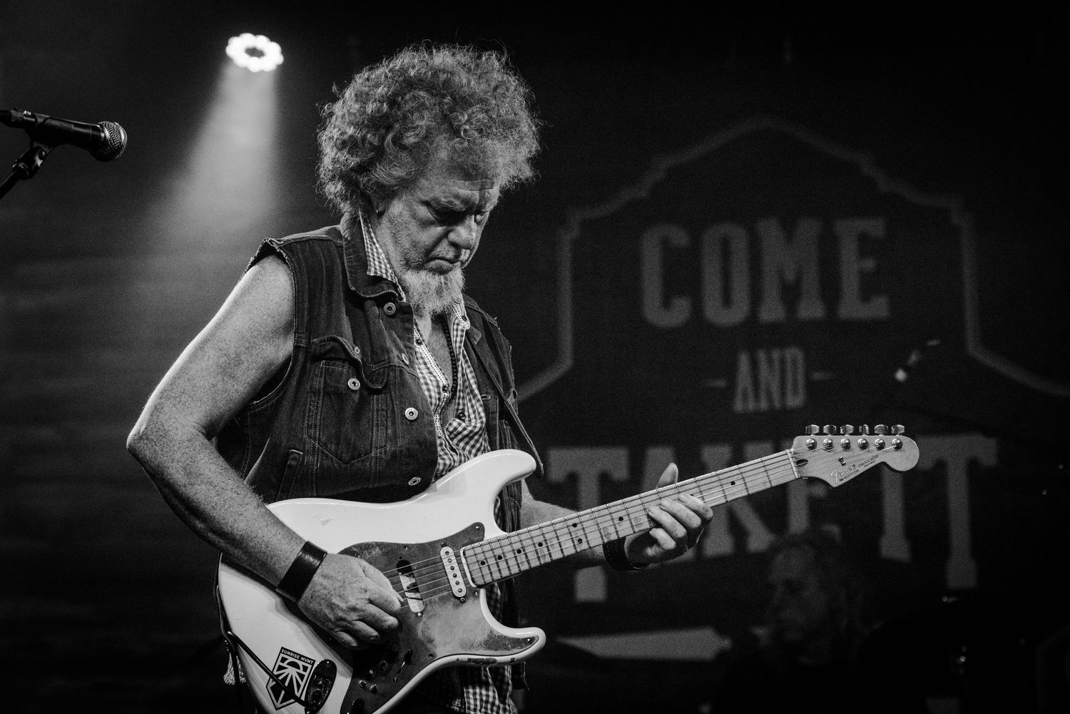 Guitarist performing on stage in black and white with curly hair and denim vest under dramatic lighting.
