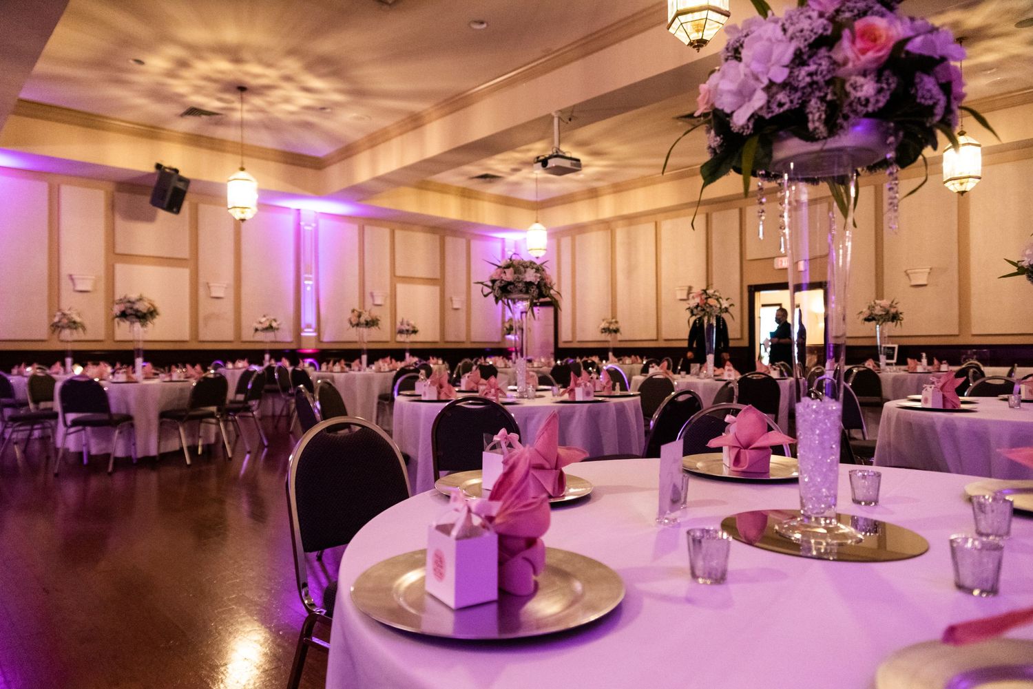 Elegant wedding reception hall decorated with purple lighting and floral centerpieces on round tables.