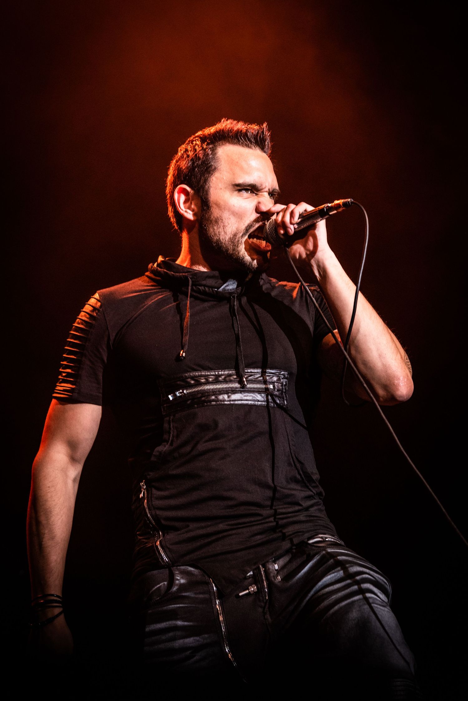 Rock singer in black tactical-style clothing performs passionately under red stage lighting.