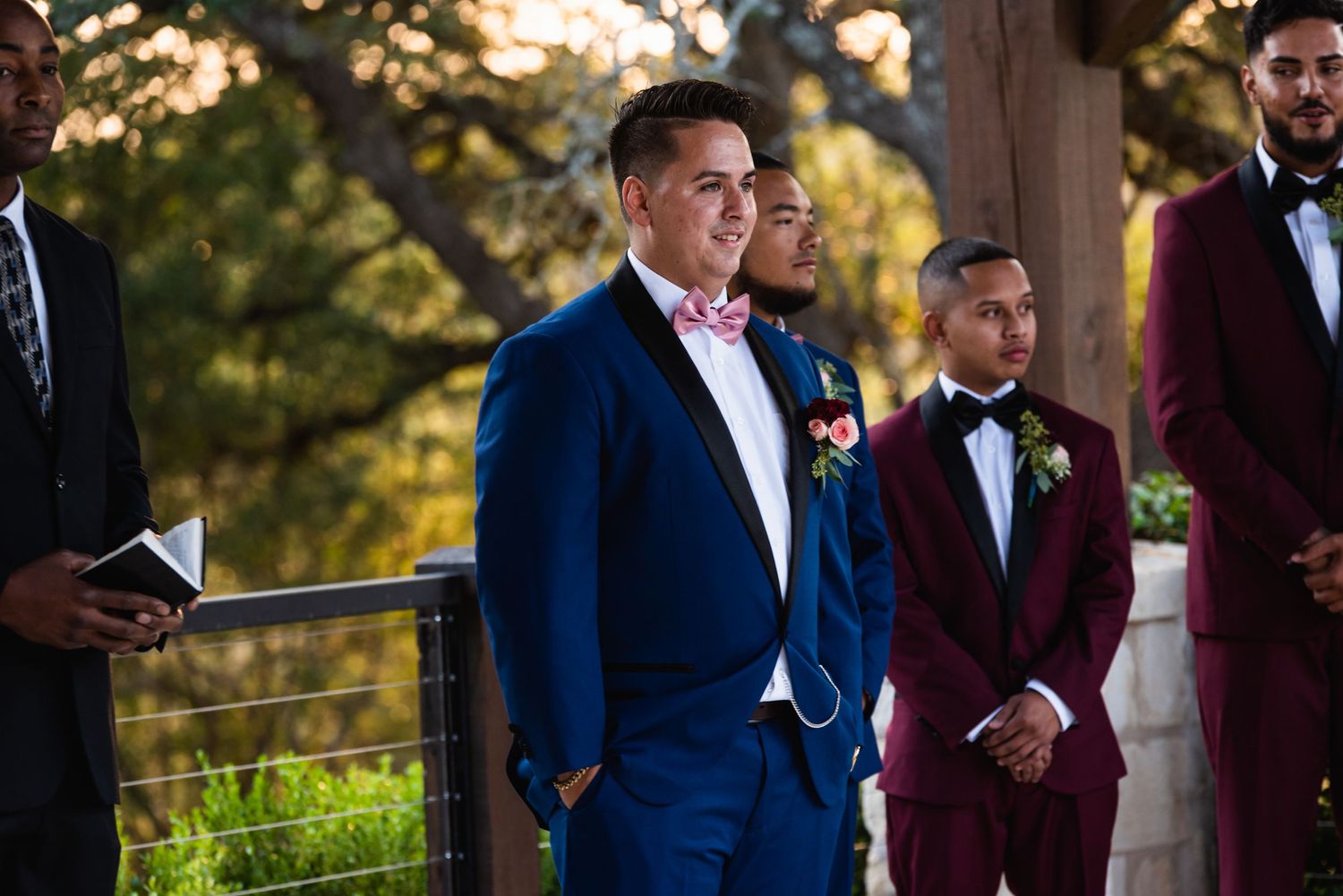 Wedding party members in blue and burgundy suits stand at outdoor ceremony.