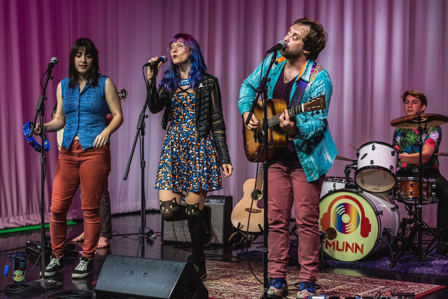 Folk band members perform on stage with guitars and drums against purple curtain.