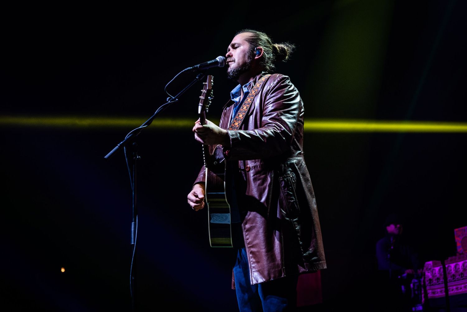 Concert performer singing passionately into microphone while playing acoustic guitar under dramatic stage lights.