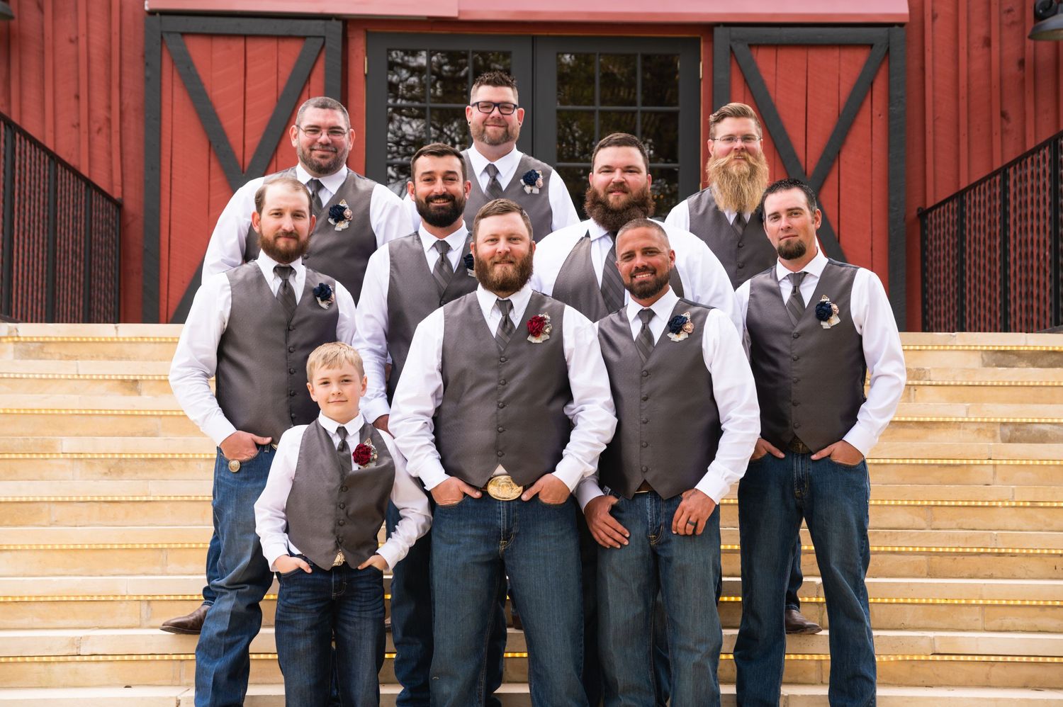 Wedding party members in matching grey vests and jeans pose on rustic red barn steps.