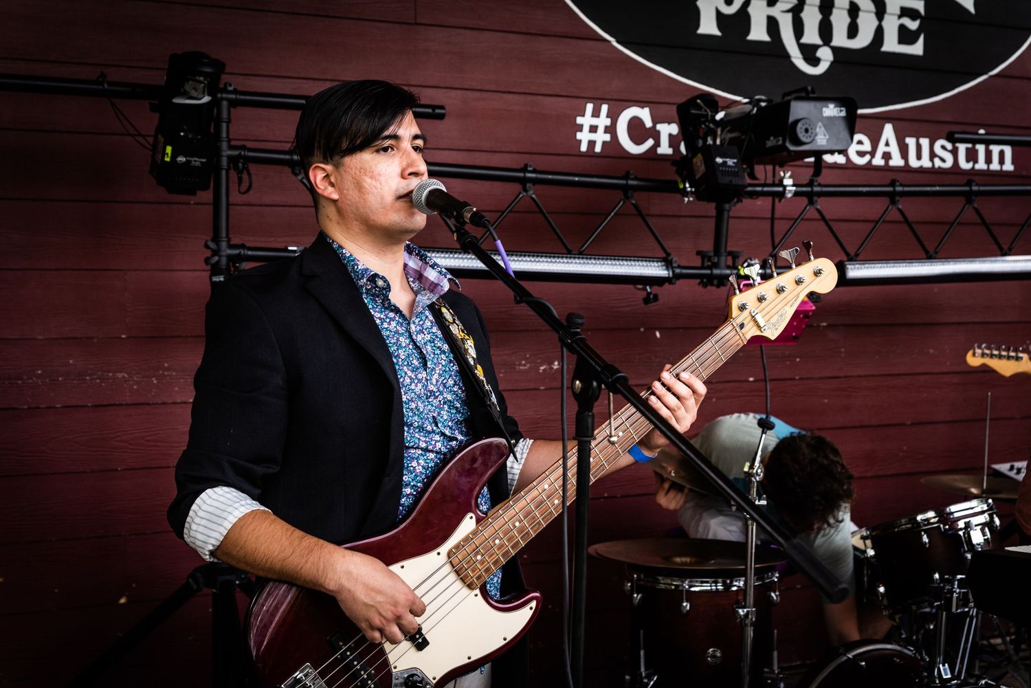 Bass player in blue patterned shirt and black blazer performs at outdoor music venue.