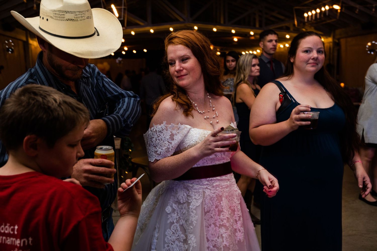 Wedding reception guests dancing and socializing under string lights with someone wearing a cowboy hat.