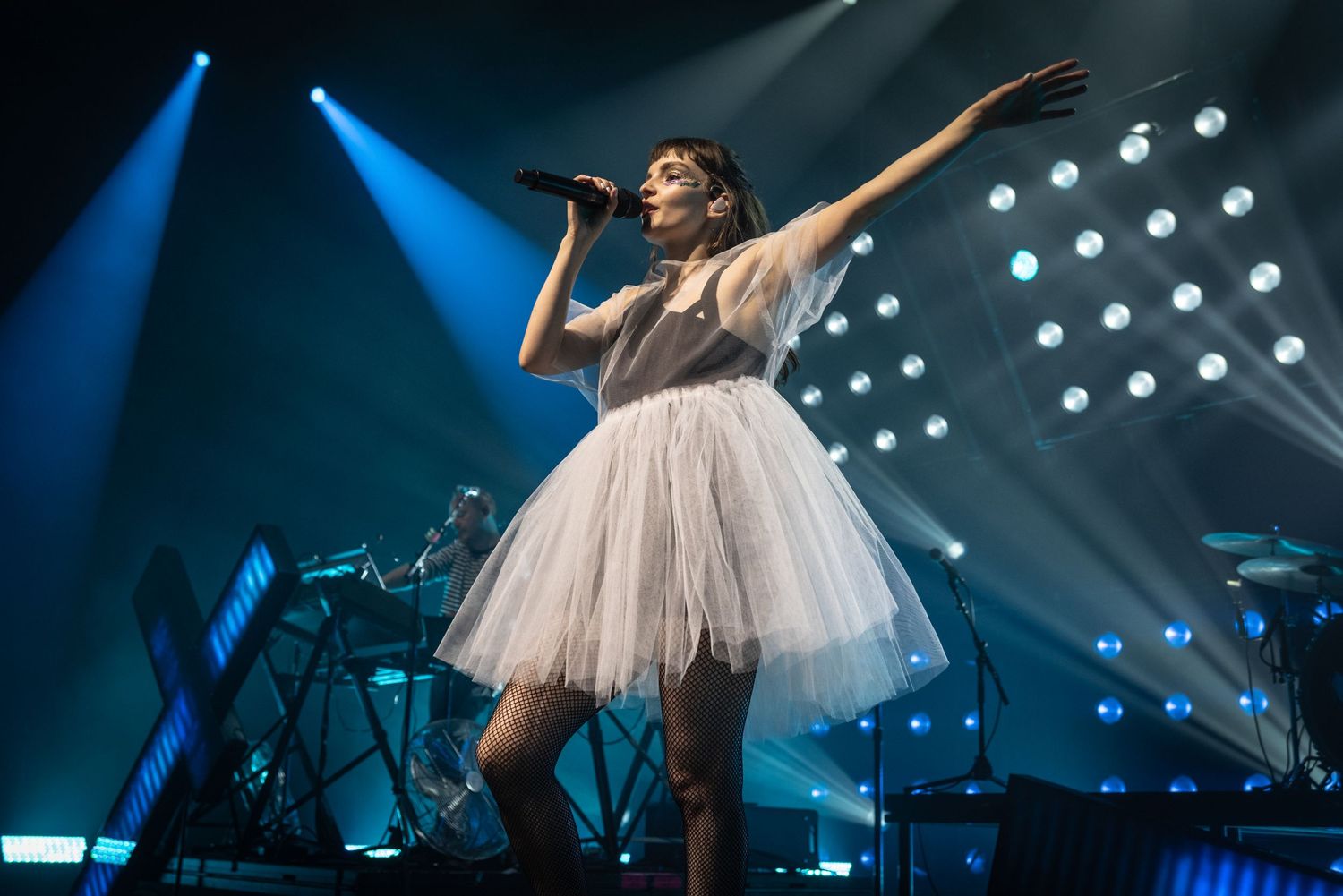 Singer in white tulle dress performs energetically under blue stage lights and spotlights.