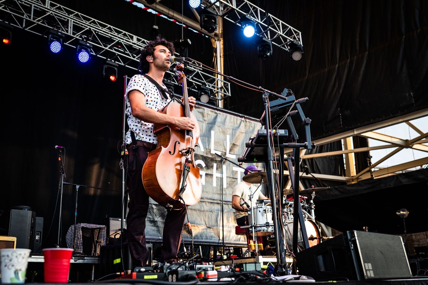 Concert performer plays acoustic guitar on festival stage with lighting equipment visible.