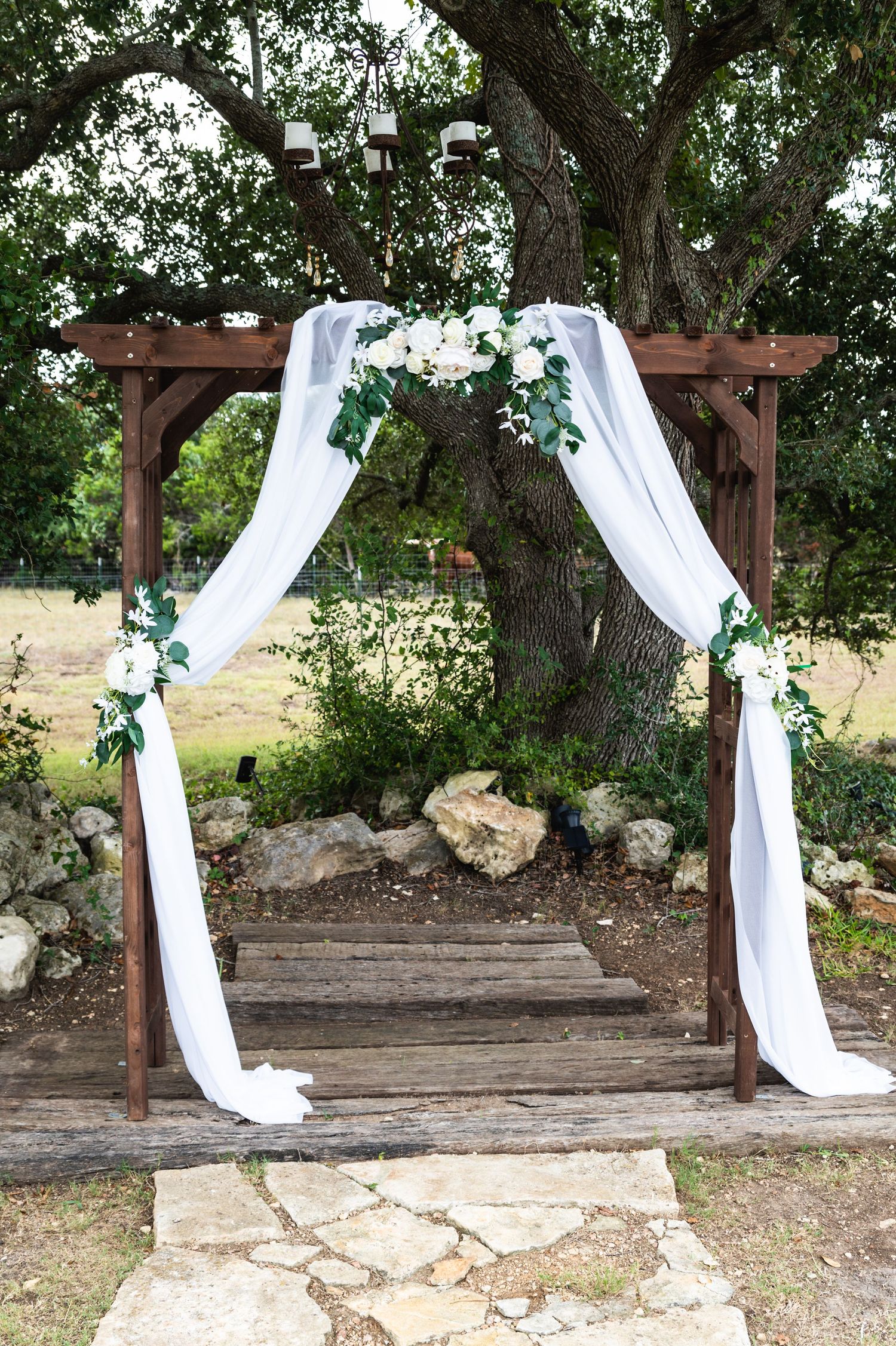 Outdoor wedding arch with white draping fabric and floral arrangements against large tree.