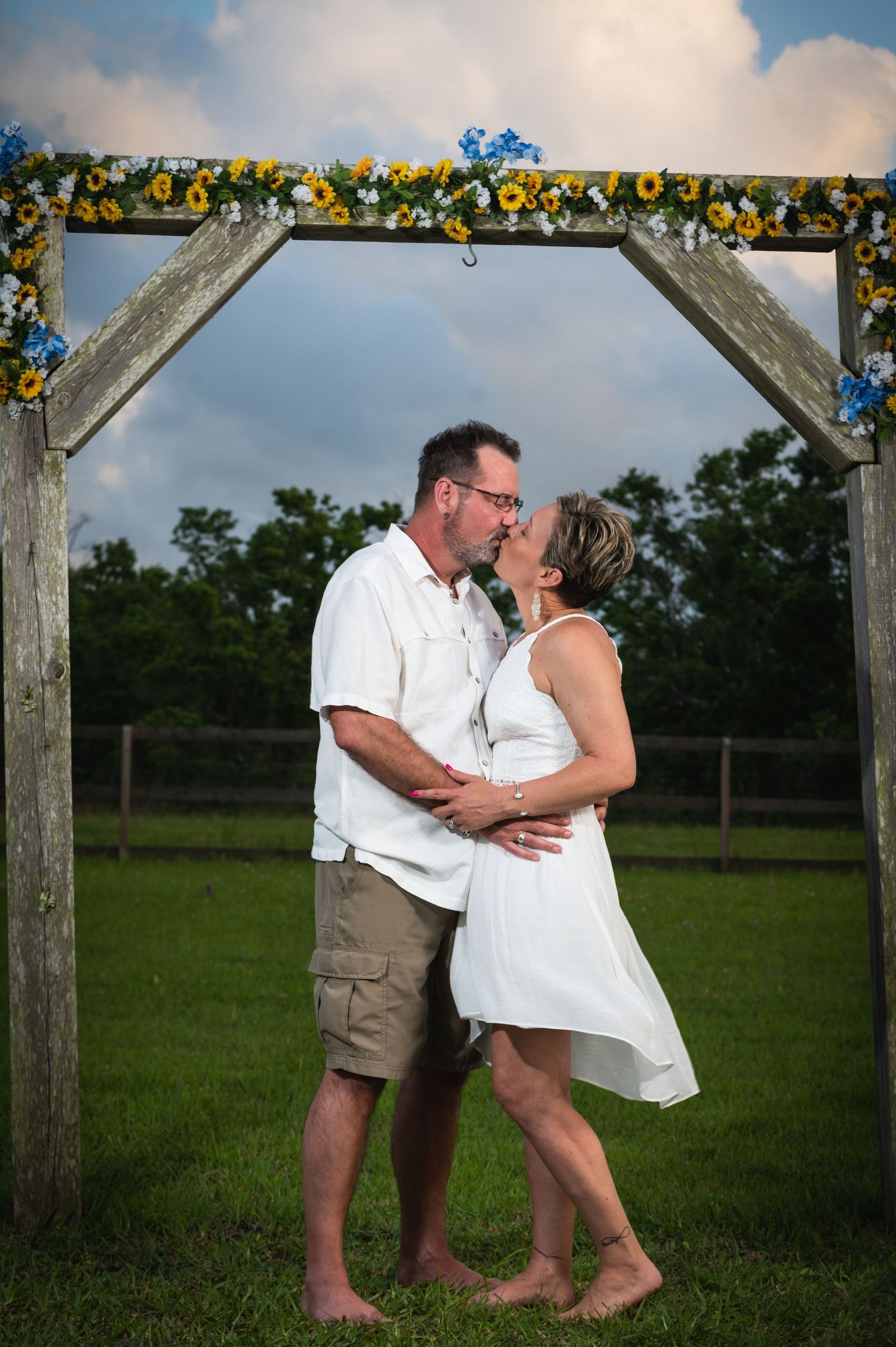 Couple sharing romantic moment under wooden arch decorated with sunflowers in outdoor setting.