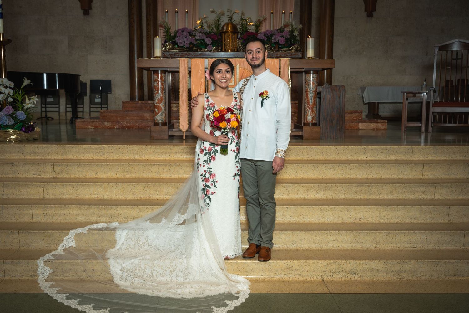 Couple stands on church steps with flowing white lace train and floral decorations.