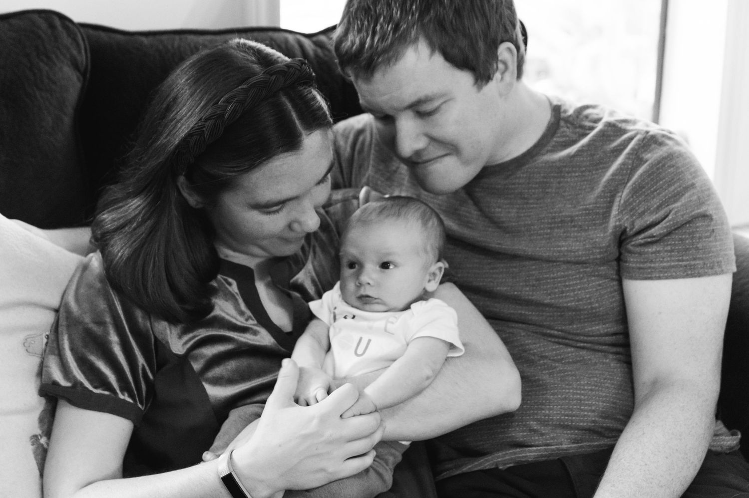 Black and white portrait of parents sharing intimate moment with newborn on couch.