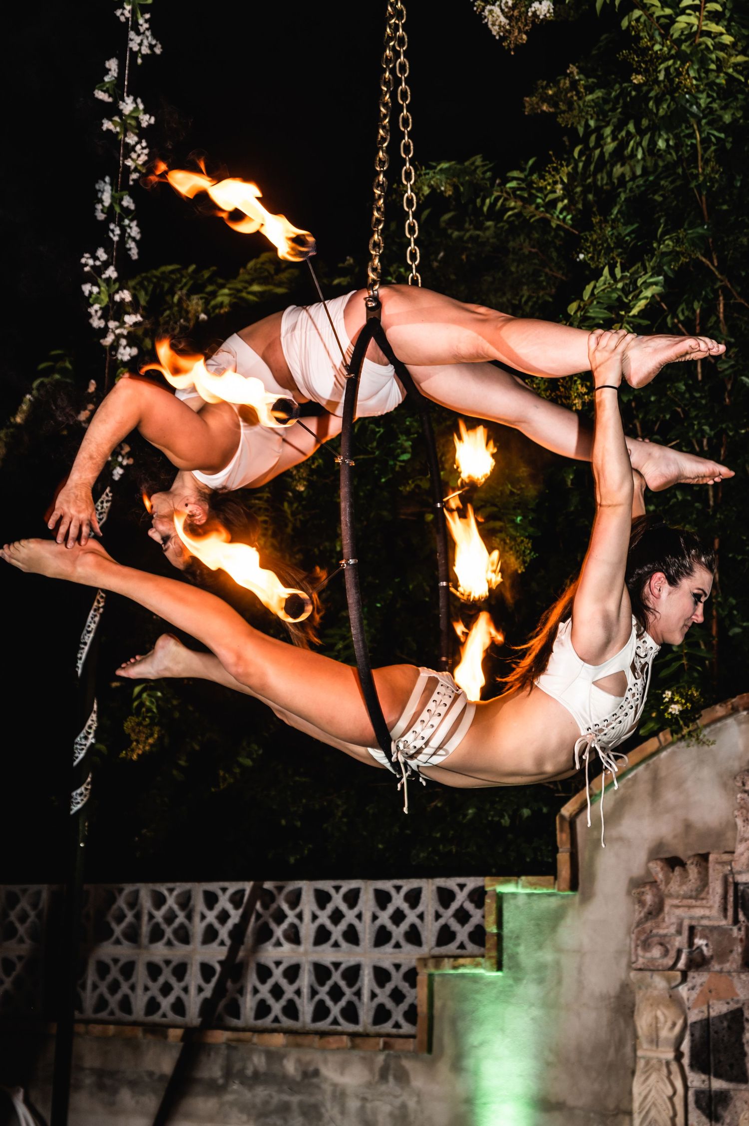 Aerial duo performs dramatic fire dance routine suspended from chains at night.
