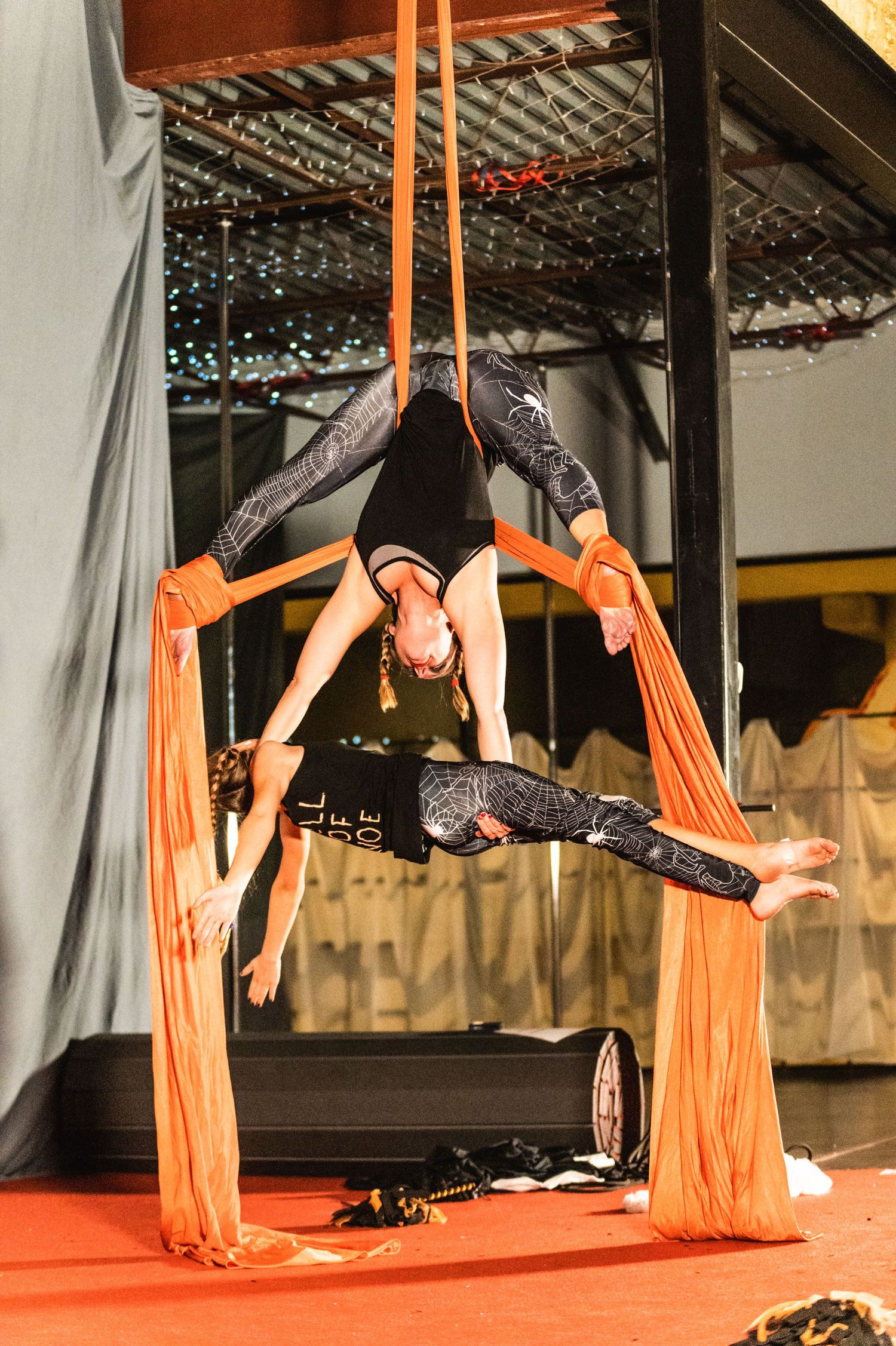 Acrobat performs split pose between orange aerial silks against sparkly backdrop.