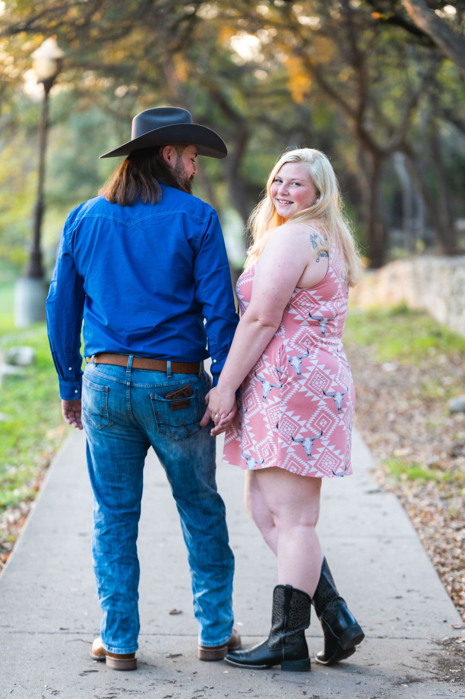 A couple in western attire poses on a tree-lined path, with him in blue and her in a pink patterned dress.