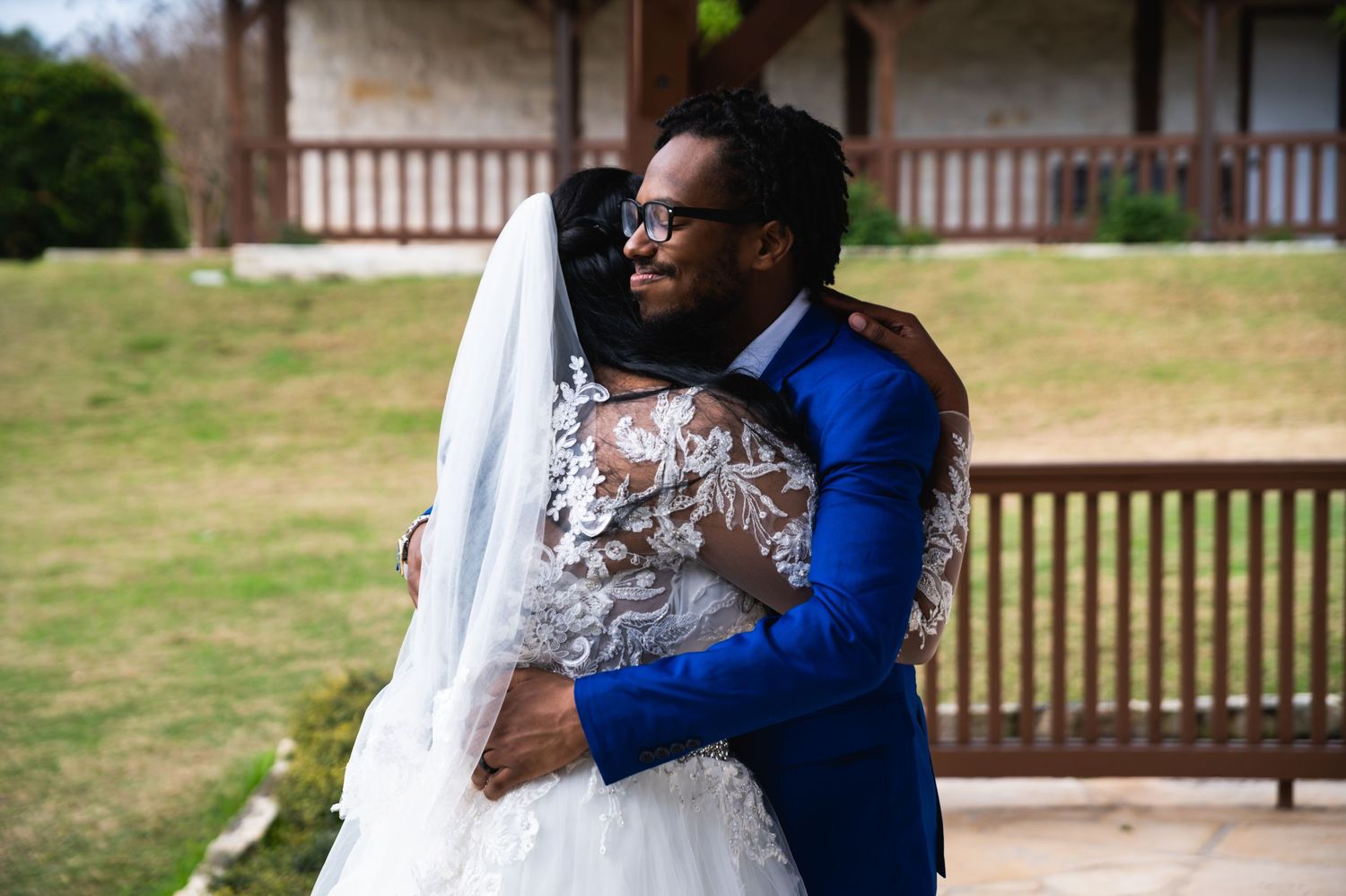 A tender embrace between newlyweds wearing a white lace gown and royal blue suit at an outdoor venue.