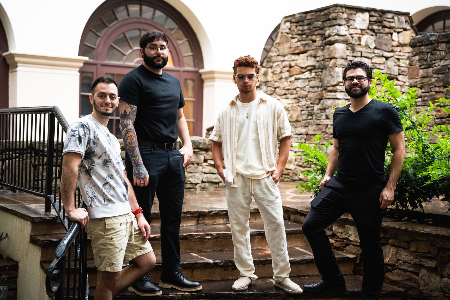 Four band members pose casually on stone steps outside a historic building with an arched doorway.