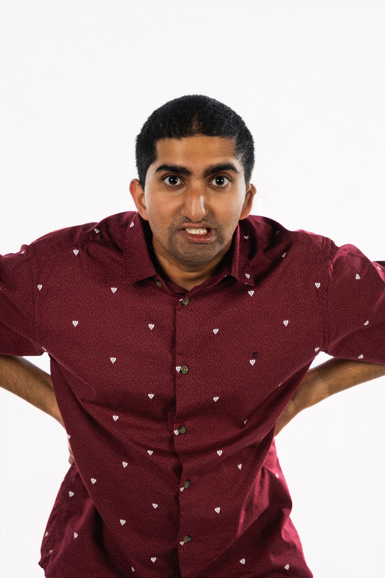 Man in burgundy heart-patterned shirt making expressive pose against white background.