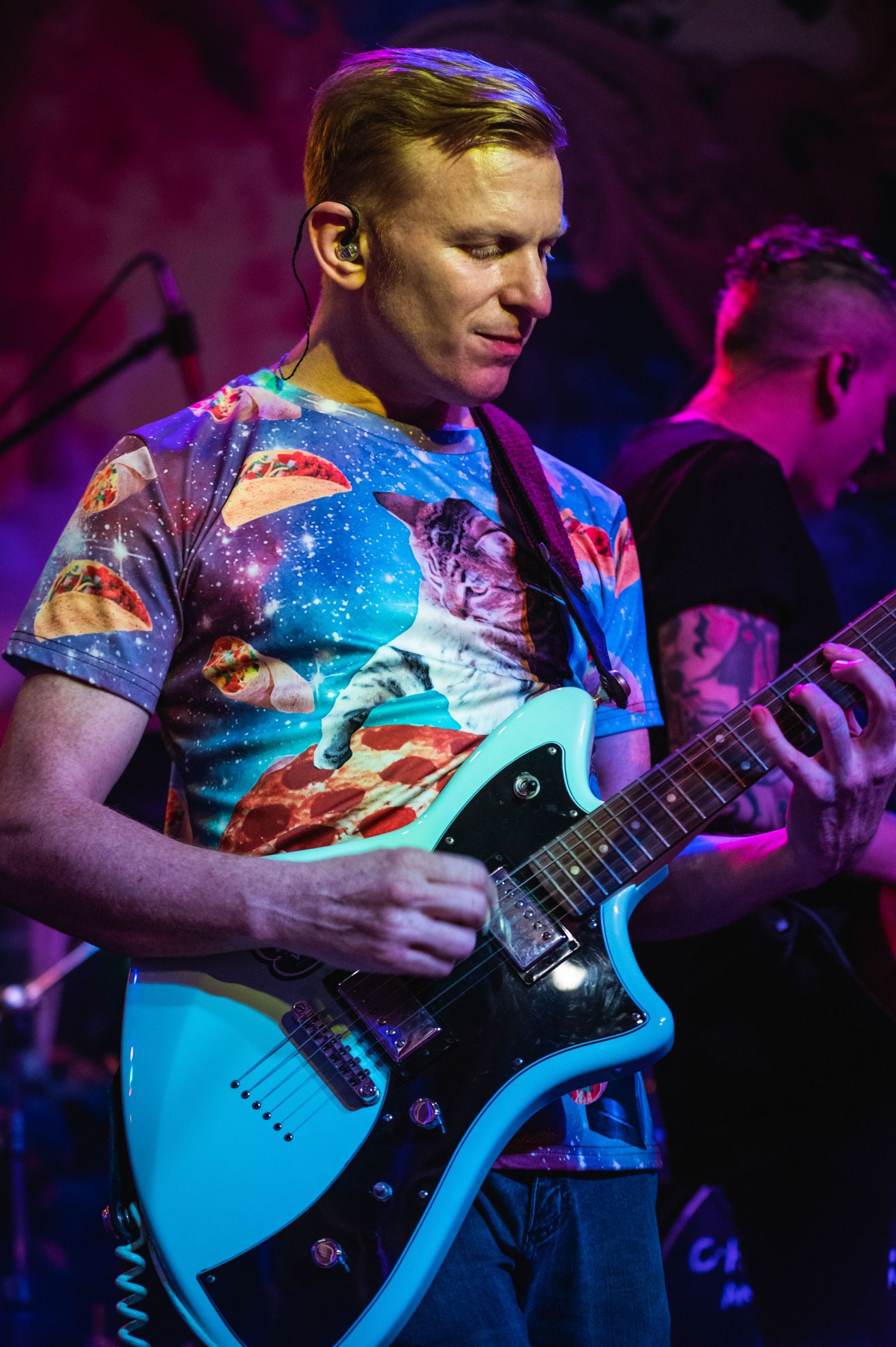Guitarist performs wearing a colorful galaxy-print t-shirt under vibrant stage lighting.