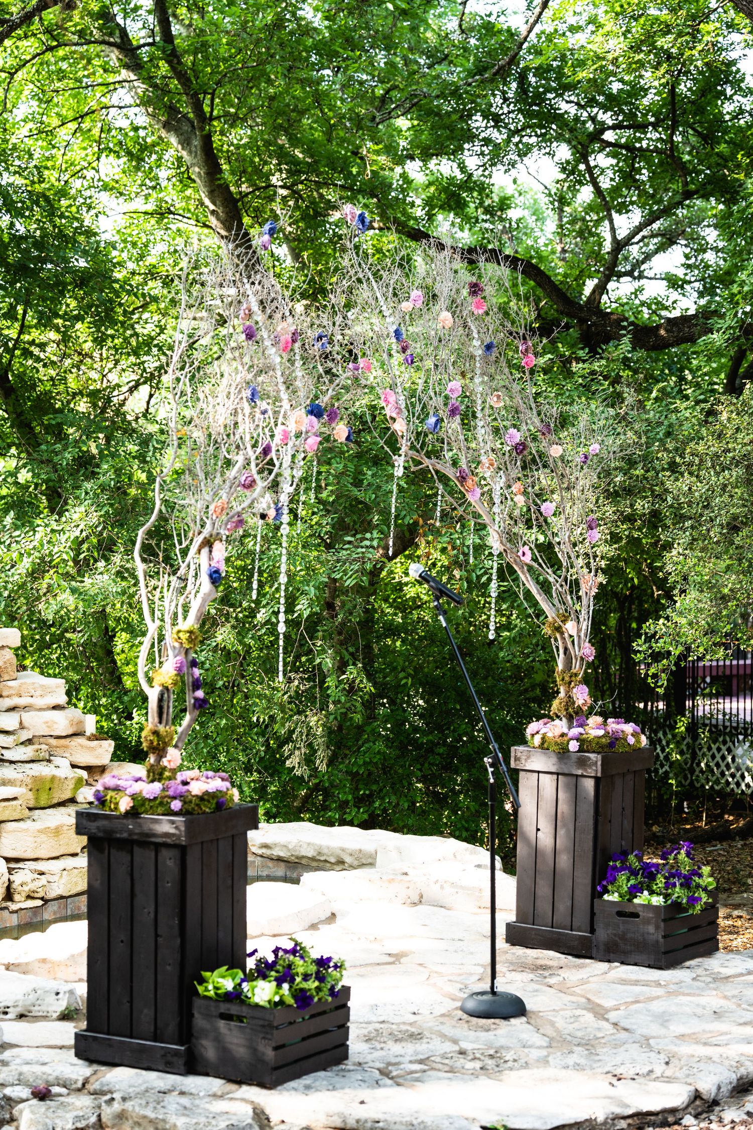 A wedding arch made of branches with purple flowers stands between wooden planters on a stone patio.