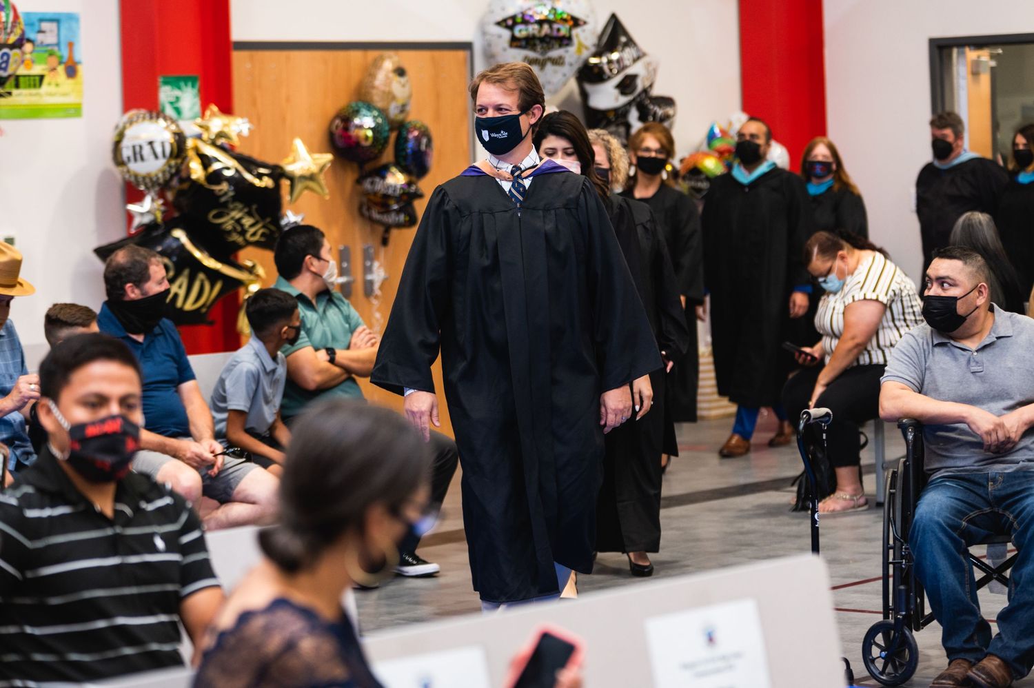 Graduate in black gown walks during ceremony while masked attendees watch from seats.