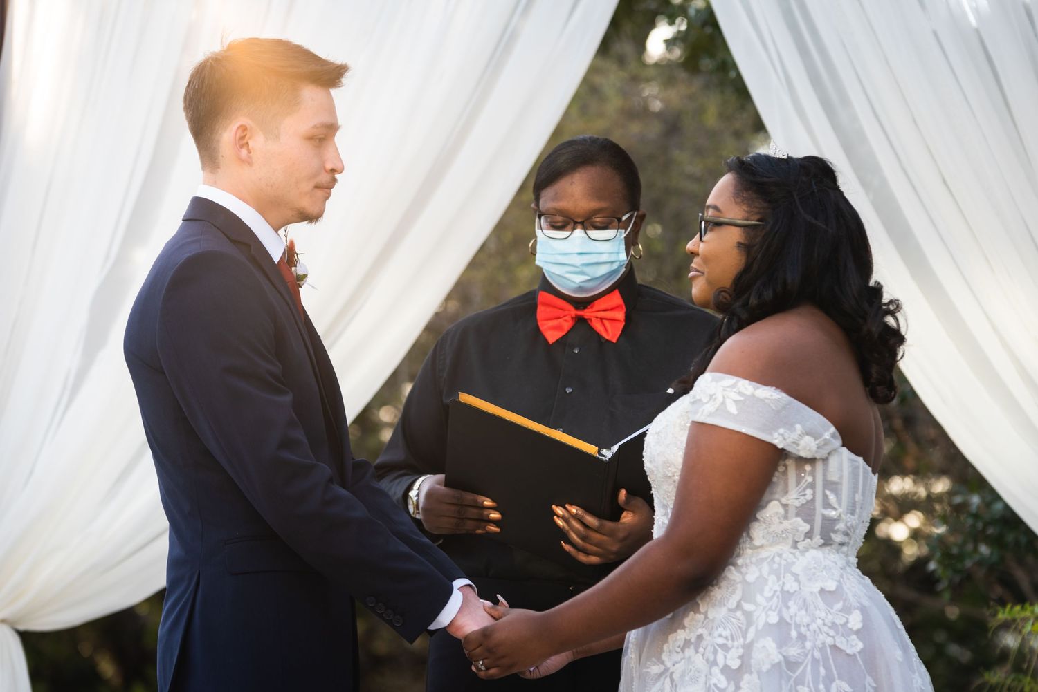 A wedding ceremony under white drapes with the officiant holding a book and wearing a red bowtie.