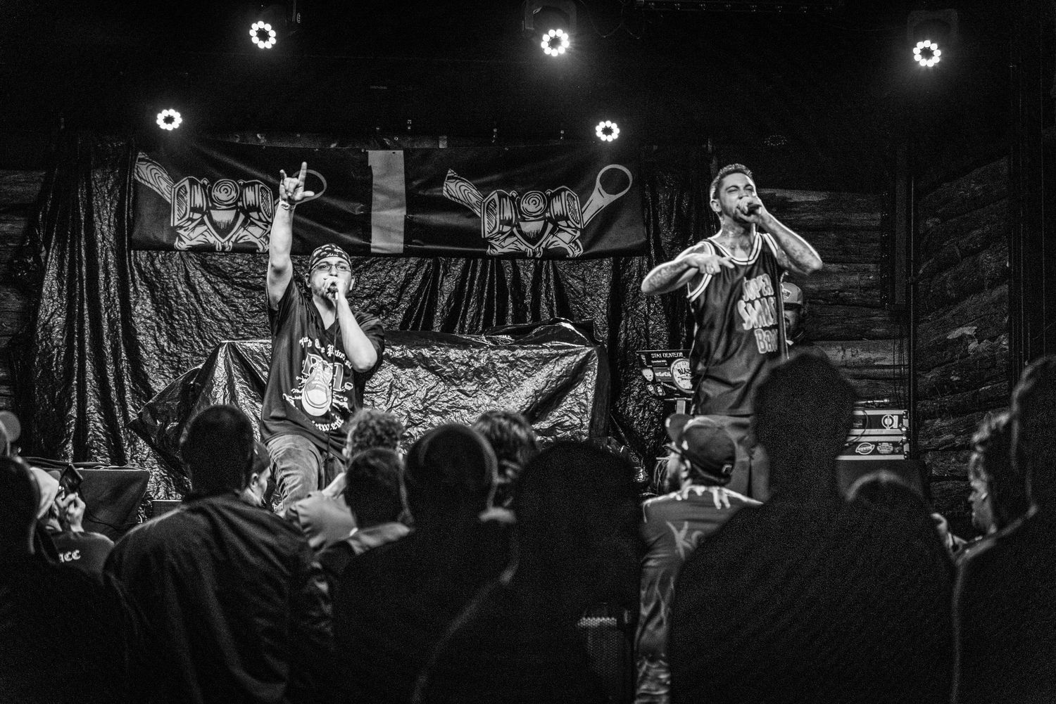 Black and white photo of performers on stage with crowd watching under circular lights.