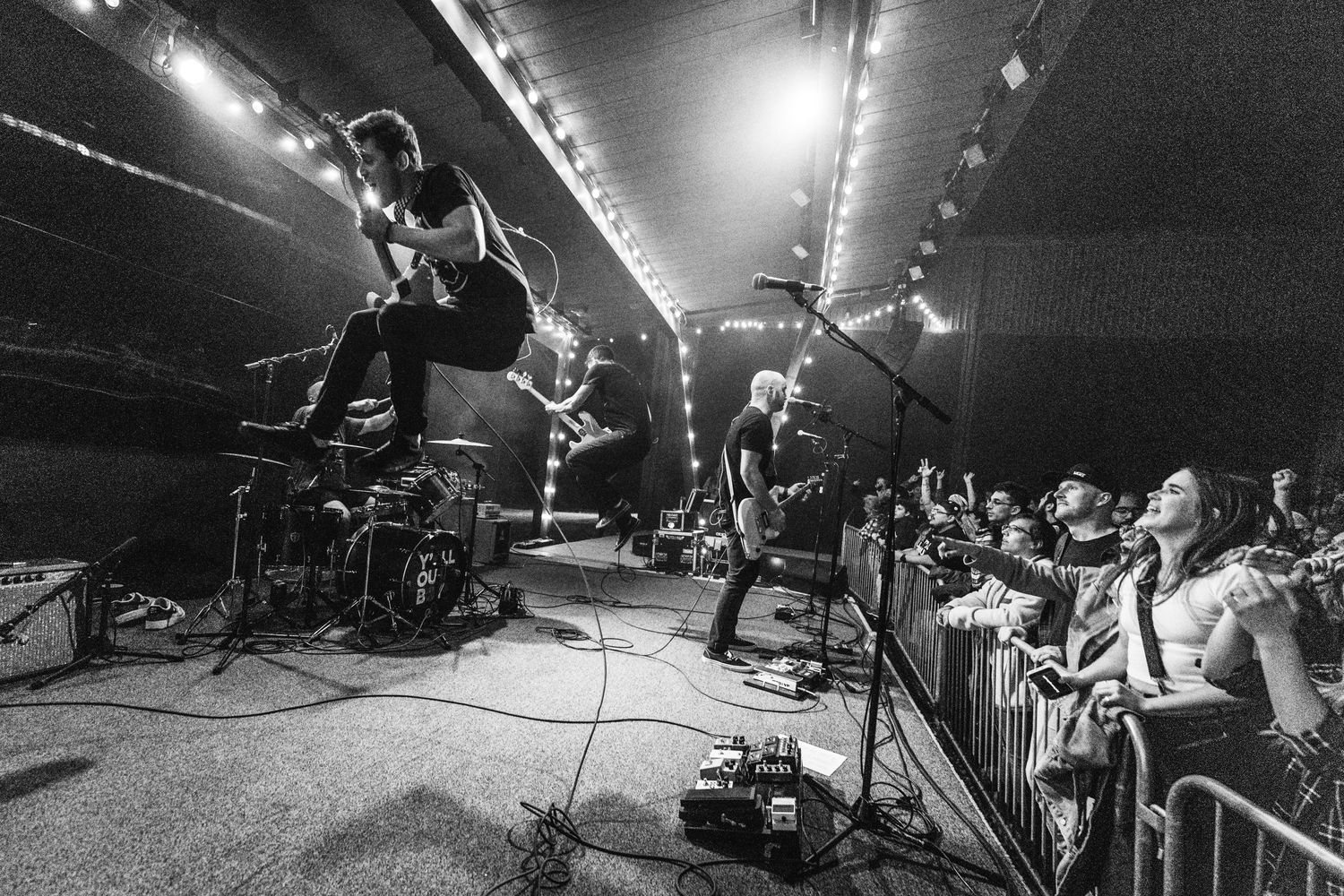 Black and white photo of energetic band performing on stage with crowd in foreground.