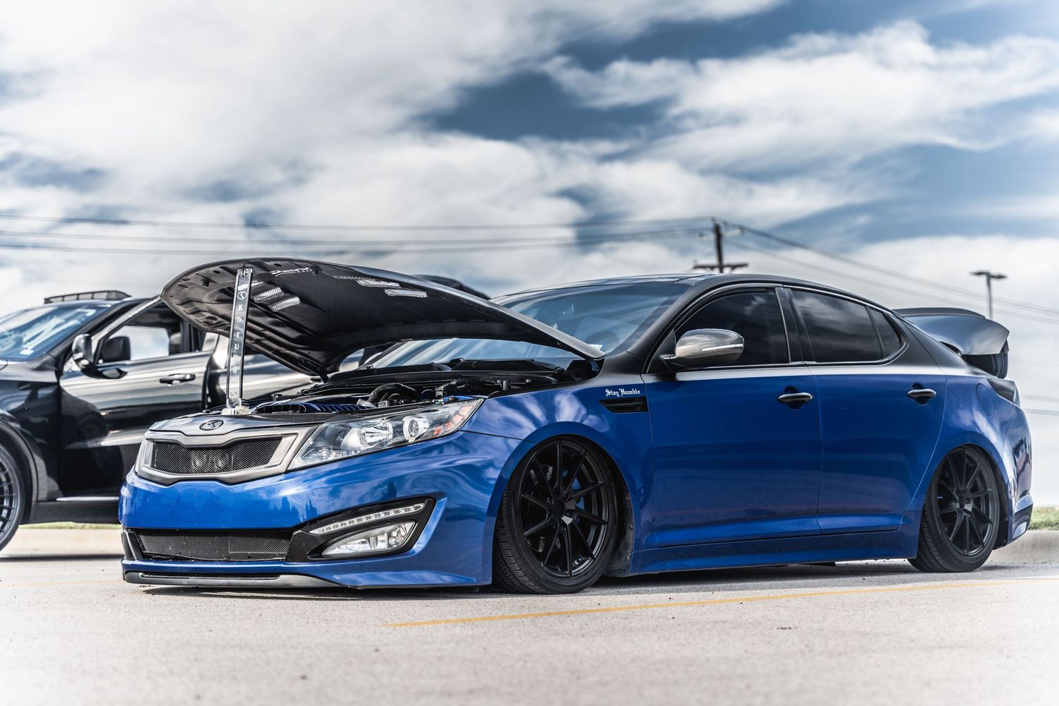 Modified blue Kia Optima with custom wheels and lowered suspension at car meet.