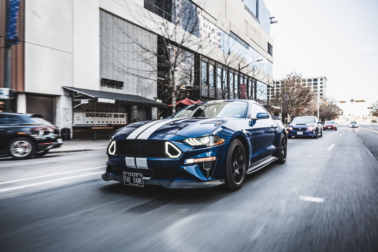 Modified Ford Mustang with blue paint and black racing stripes photographed in motion on urban street.