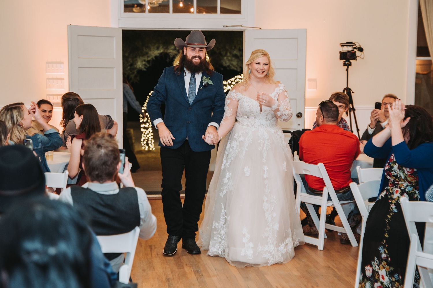 A couple walks down the aisle while wedding guests applaud in a bright indoor reception space.
