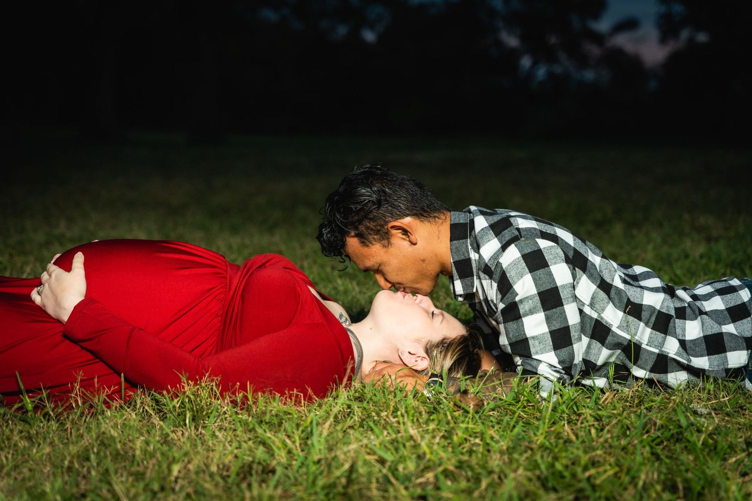 A couple in romantic pose lying on grass with sunset lighting in background.
