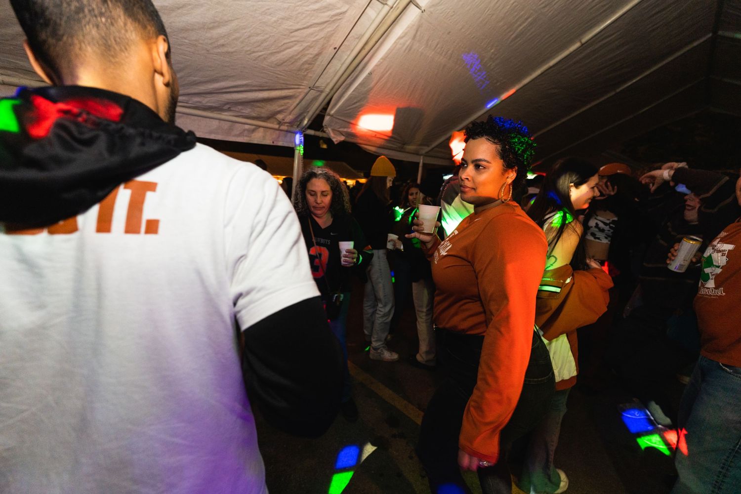 People dancing and socializing under a dark tent with colorful lights at a nighttime party event.