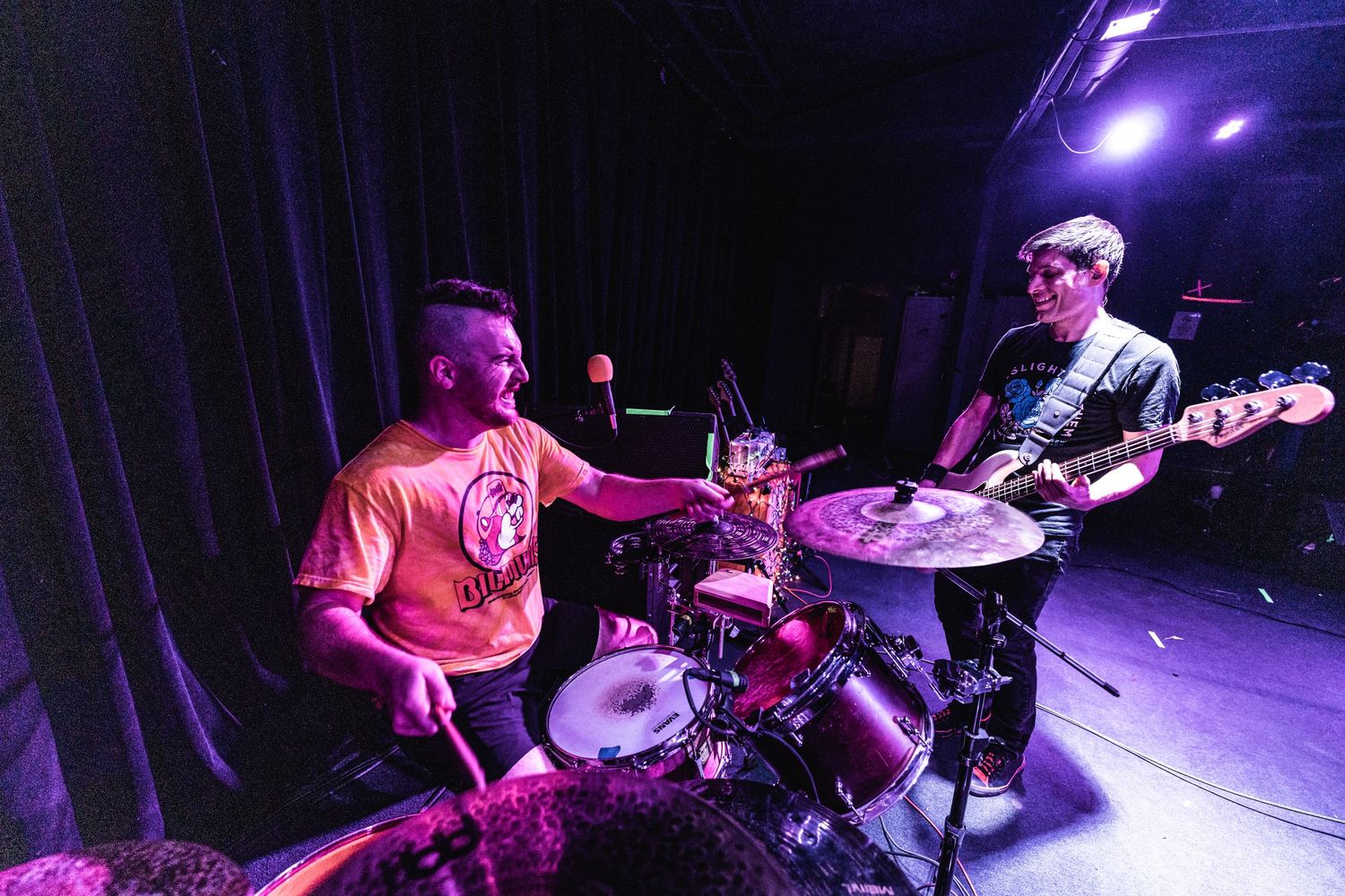 Band members perform energetically on stage under purple lighting with a drummer and bassist visible.