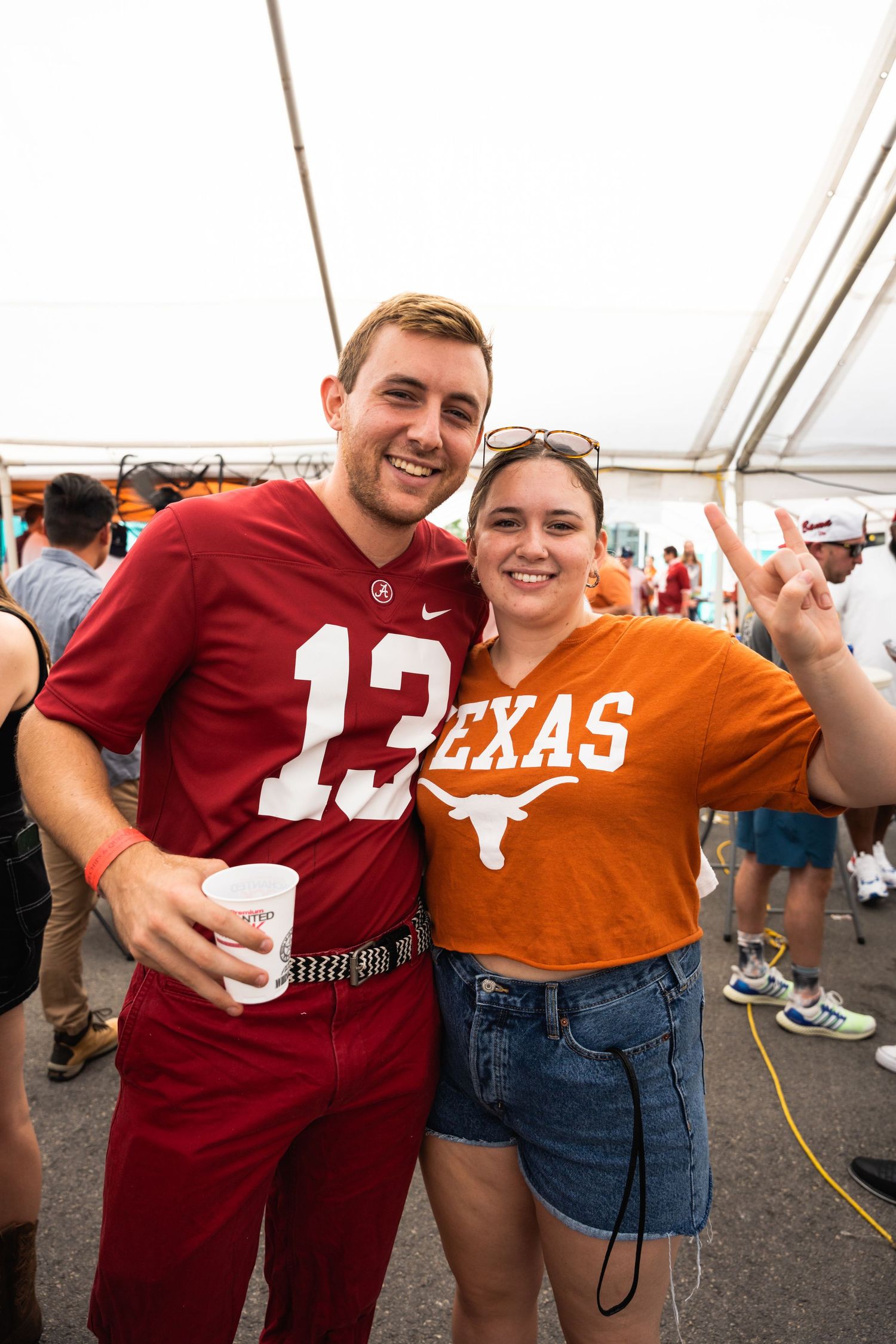 College football fans in contrasting Texas and Alabama team colors celebrate together under a tent.