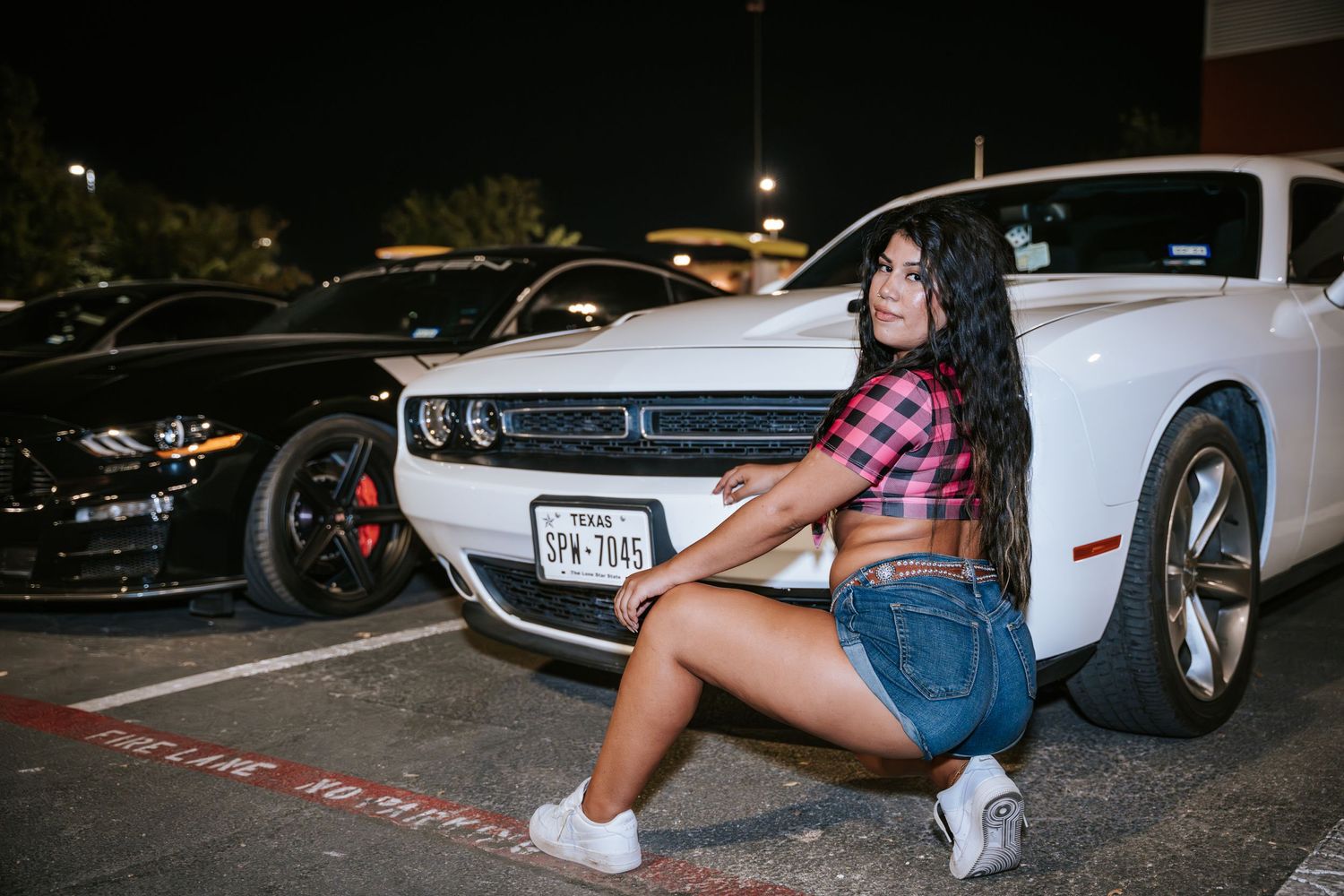 Model in pink crop top and denim shorts poses with white Dodge Challenger.