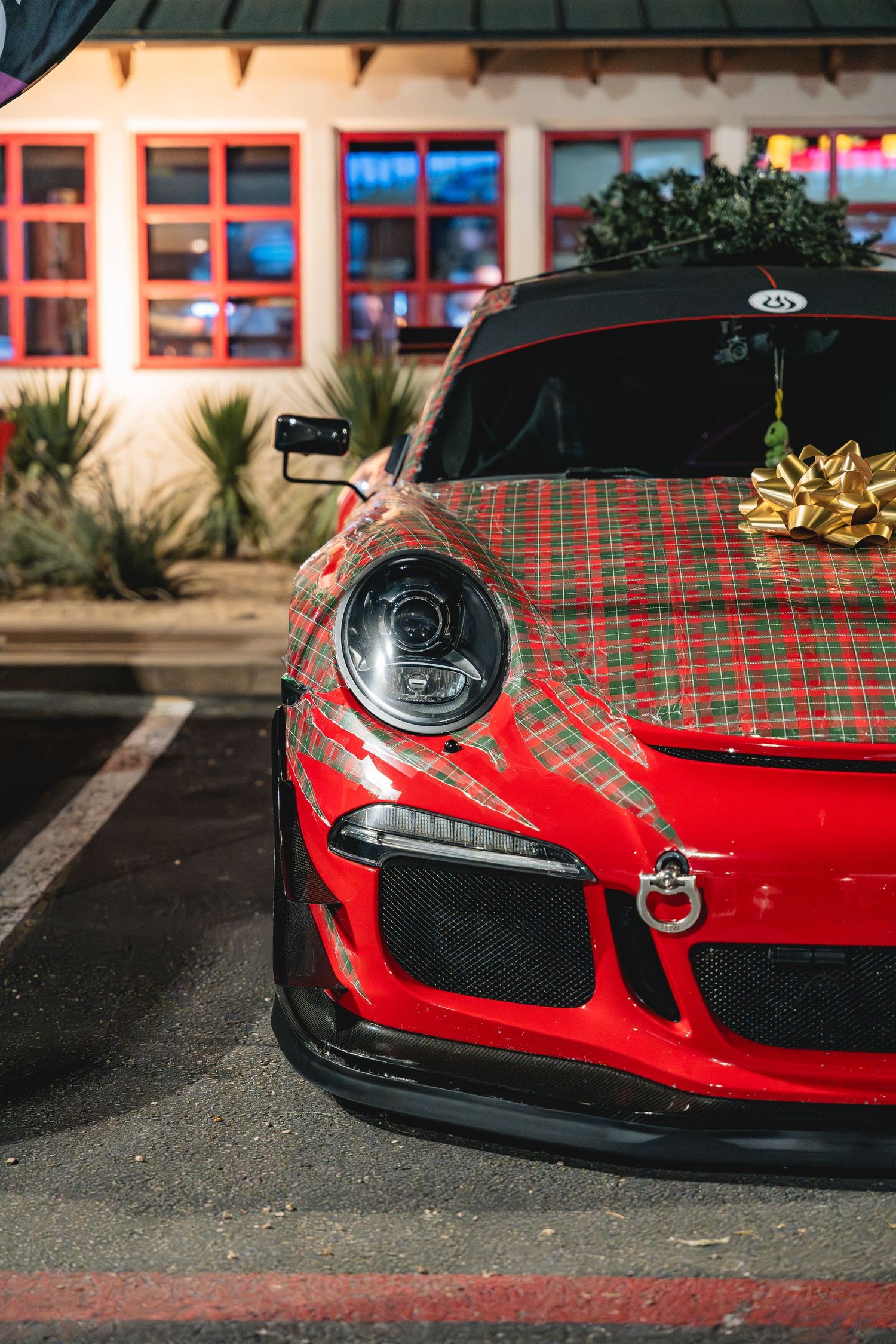 Close-up front view of a red Porsche 911 GT3 with plaid hood wrap parked outside a building.