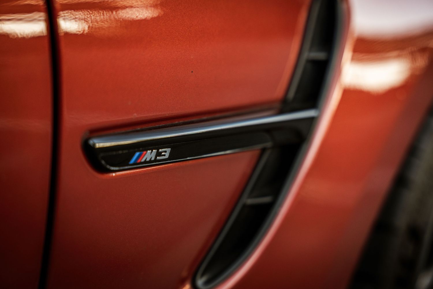 Close-up of BMW M3 side gill badge on orange metallic paintwork.