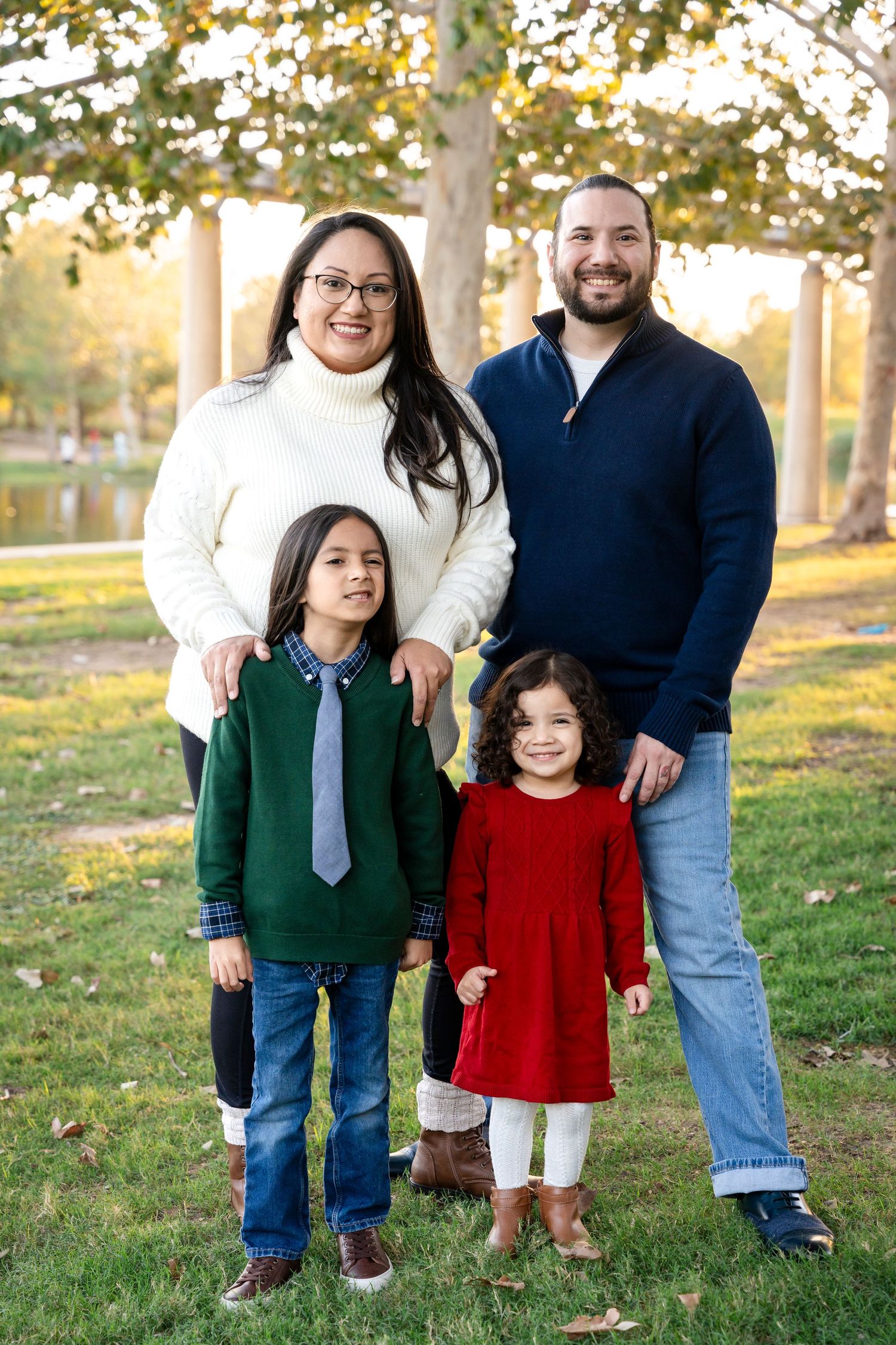 Family portrait of four standing together on grass with autumn trees in background.
