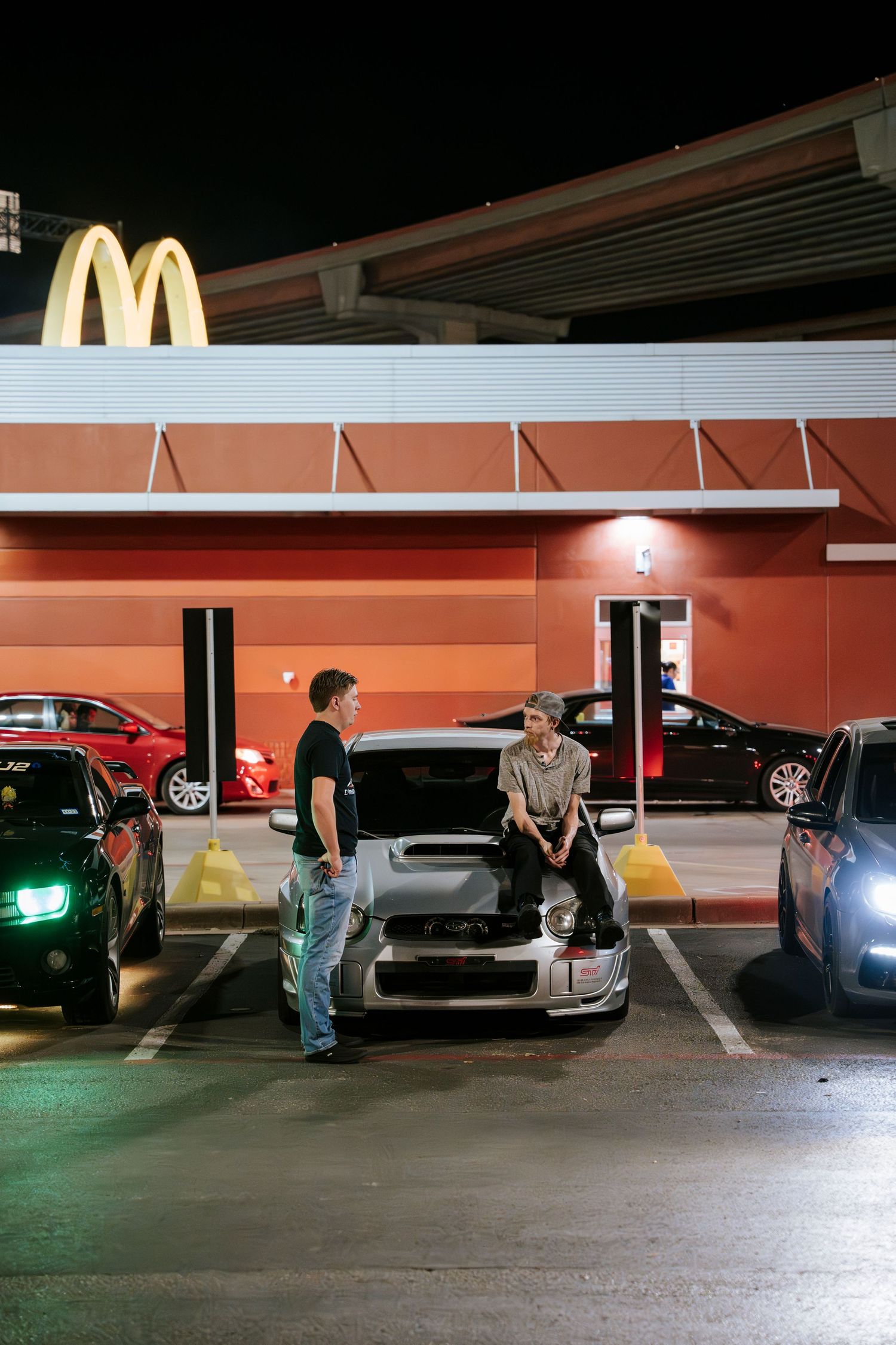 People gathered around cars in a McDonald's parking lot at night with illuminated golden arches visible.