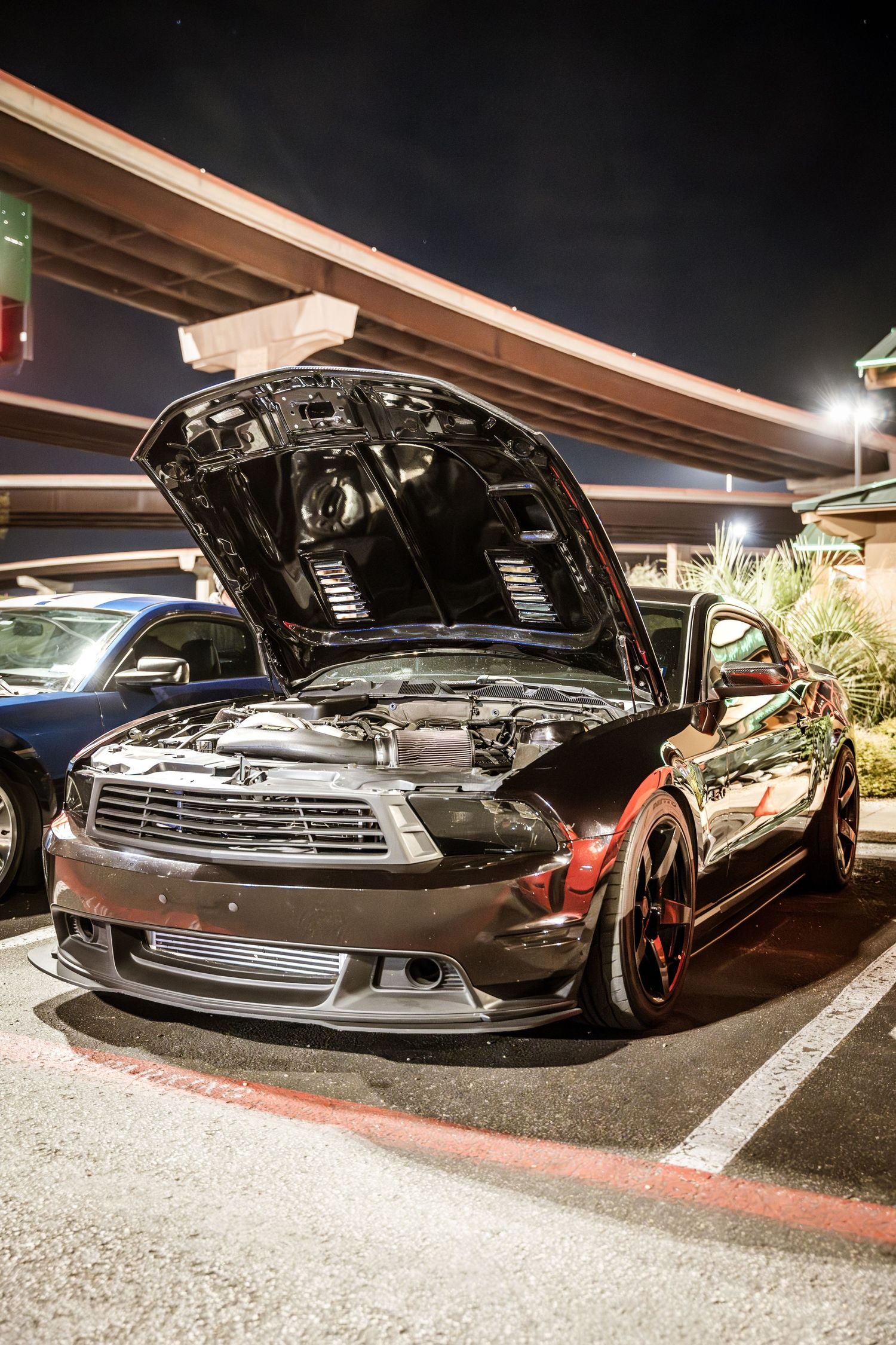 A modified black Ford Mustang with open hood displaying engine at night.