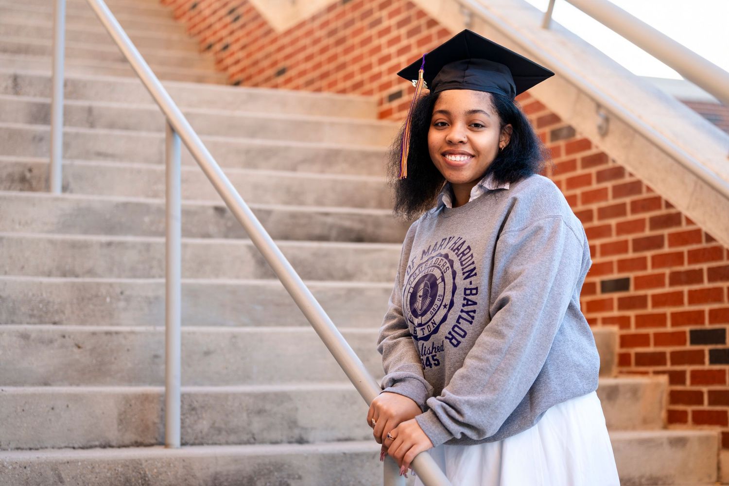 A graduate in a grey university sweatshirt and cap poses on concrete steps near brick walls.