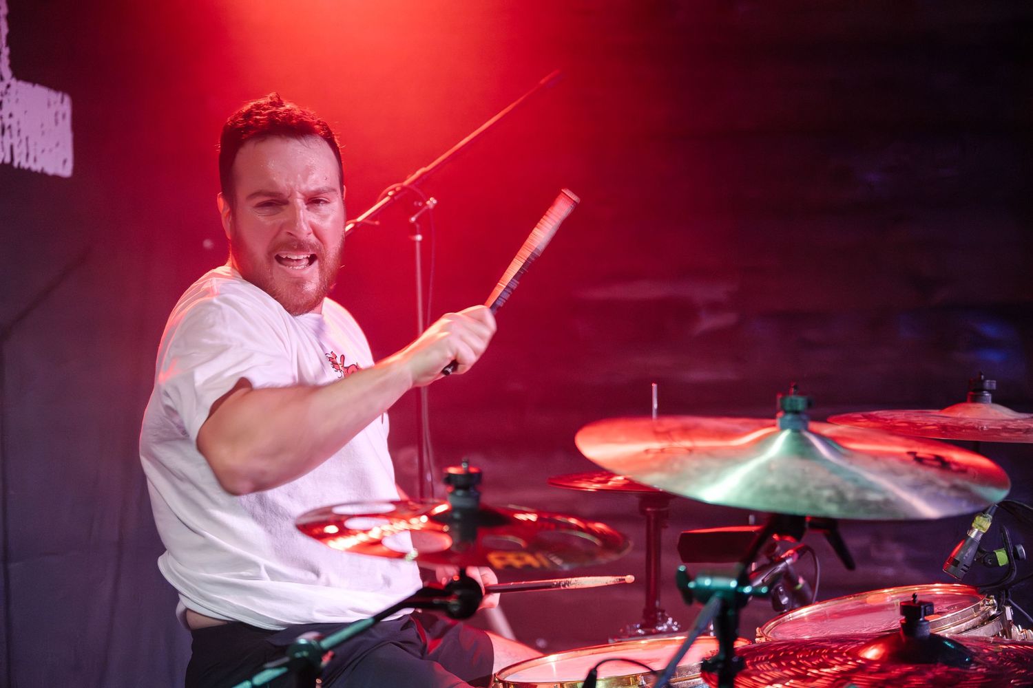Drummer performs energetically behind drum kit under red stage lighting.