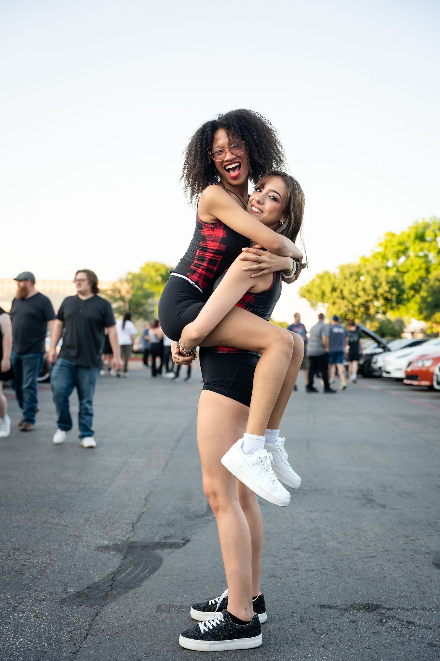 Two friends embracing and laughing together in a parking lot while wearing casual summer outfits.