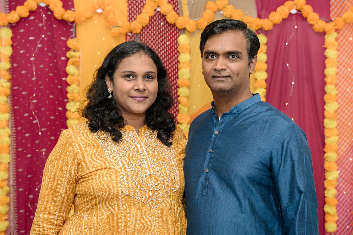Couple posing in traditional Indian clothing against colorful backdrop with orange and red decorations.