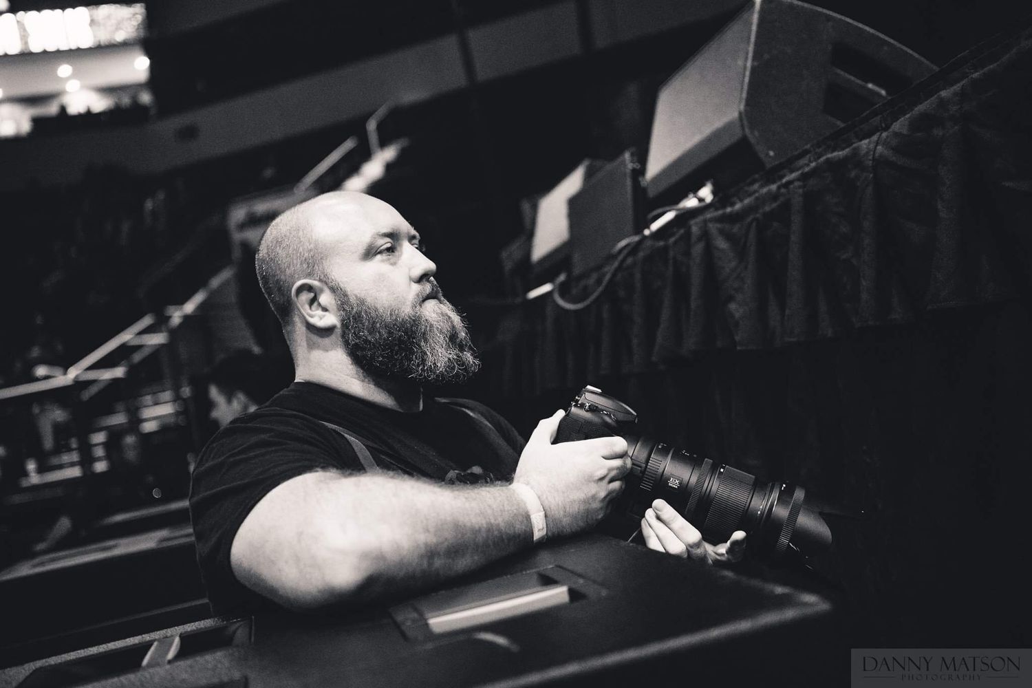 A person with a beard performs on stage with dramatic lighting and musical equipment in the background.