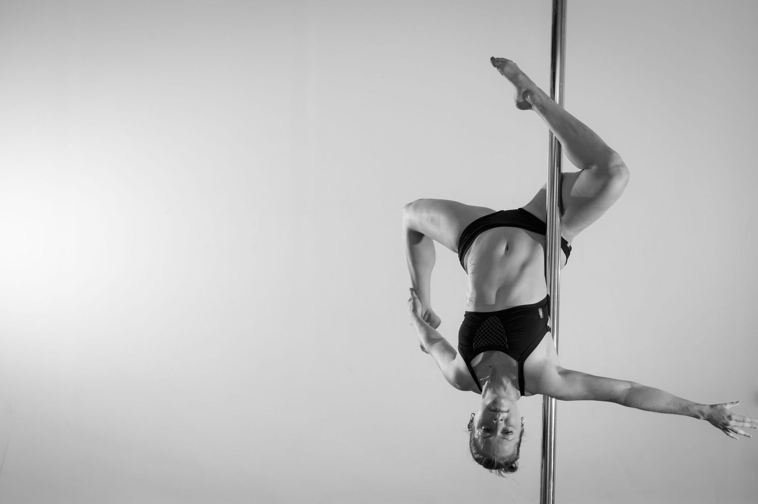 A pole dancer performs a graceful split pose on a vertical pole in a black and white artistic photograph.
