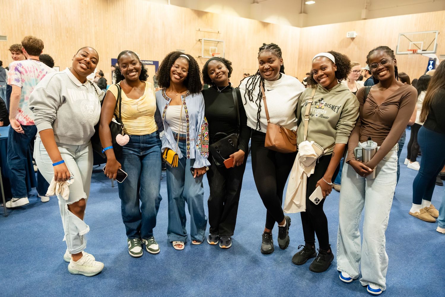 Seven young women smiling and posing together at an indoor social event on a blue carpeted floor.